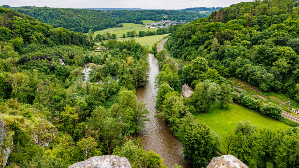 Packrafter paddling on the Ourthe surrounded by green hills and limestone cliffs, such as Roche aux Corneilles and Les rochers de Sy, during a sunny day.